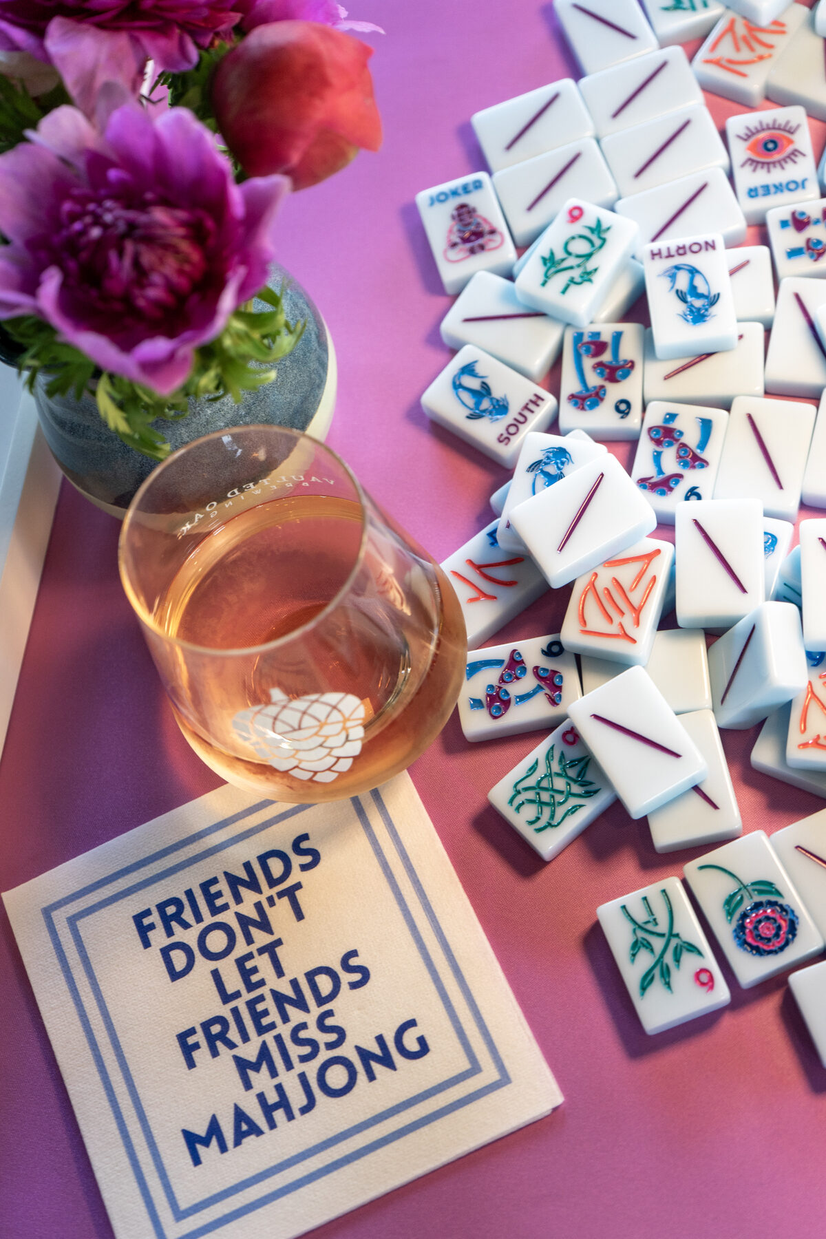 A 'Friends Don't Let Friends Miss Mahjong' card beside peonies, mahjong tiles, and a rosé